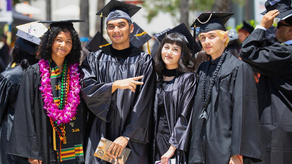 Four graduating students smile together in black caps and gowns. The person on the left wears a vibrant purple orchid lei. The student next to them gives a peace sign. They all stand close outdoors during a commencement ceremony.
