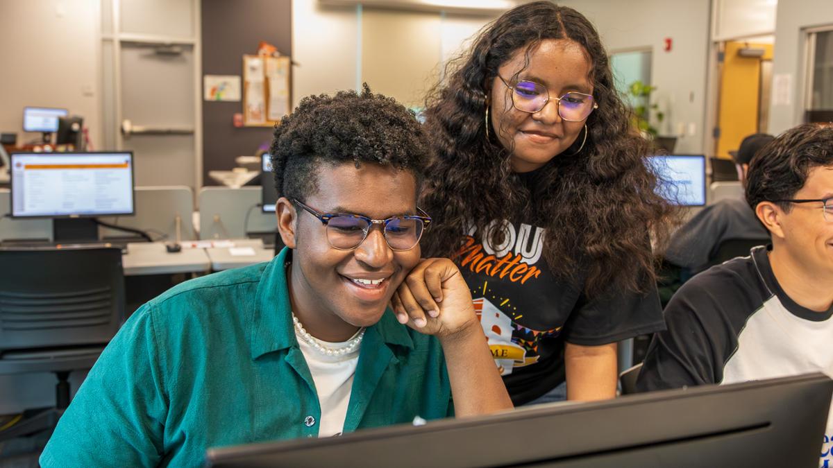 Two college students work together at a computer in a classroom lab. One student sits smiling at the screen while another stands beside them looking on. Other students and computer stations are visible in the background.