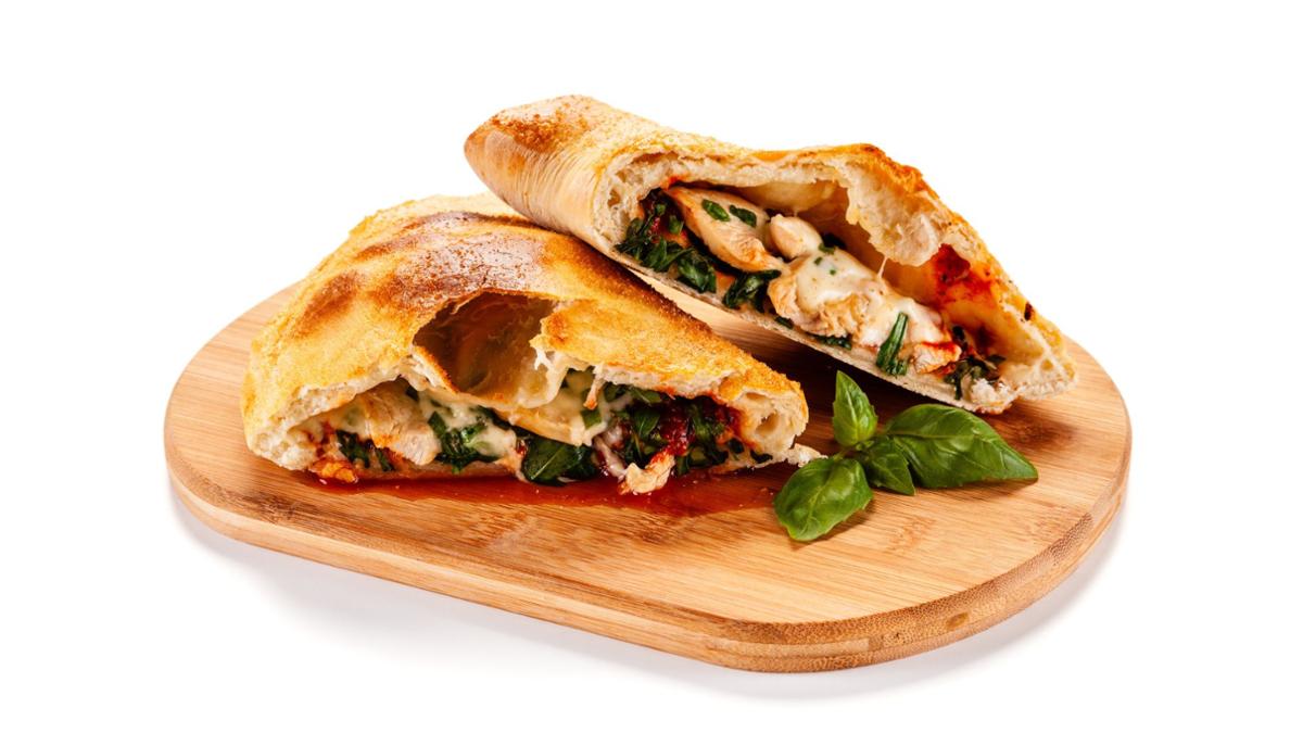A golden-brown calzone cut in half on a wooden board, filled with melted cheese, spinach, chicken, and tomato sauce, with fresh basil leaves beside it.